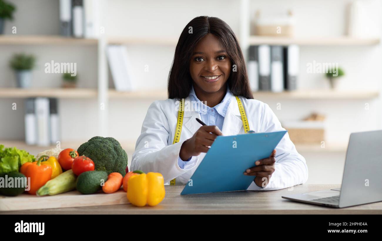 Positive female african american nutritionist with clipboard creating plant based meal plan for patient, panorama Stock Photo