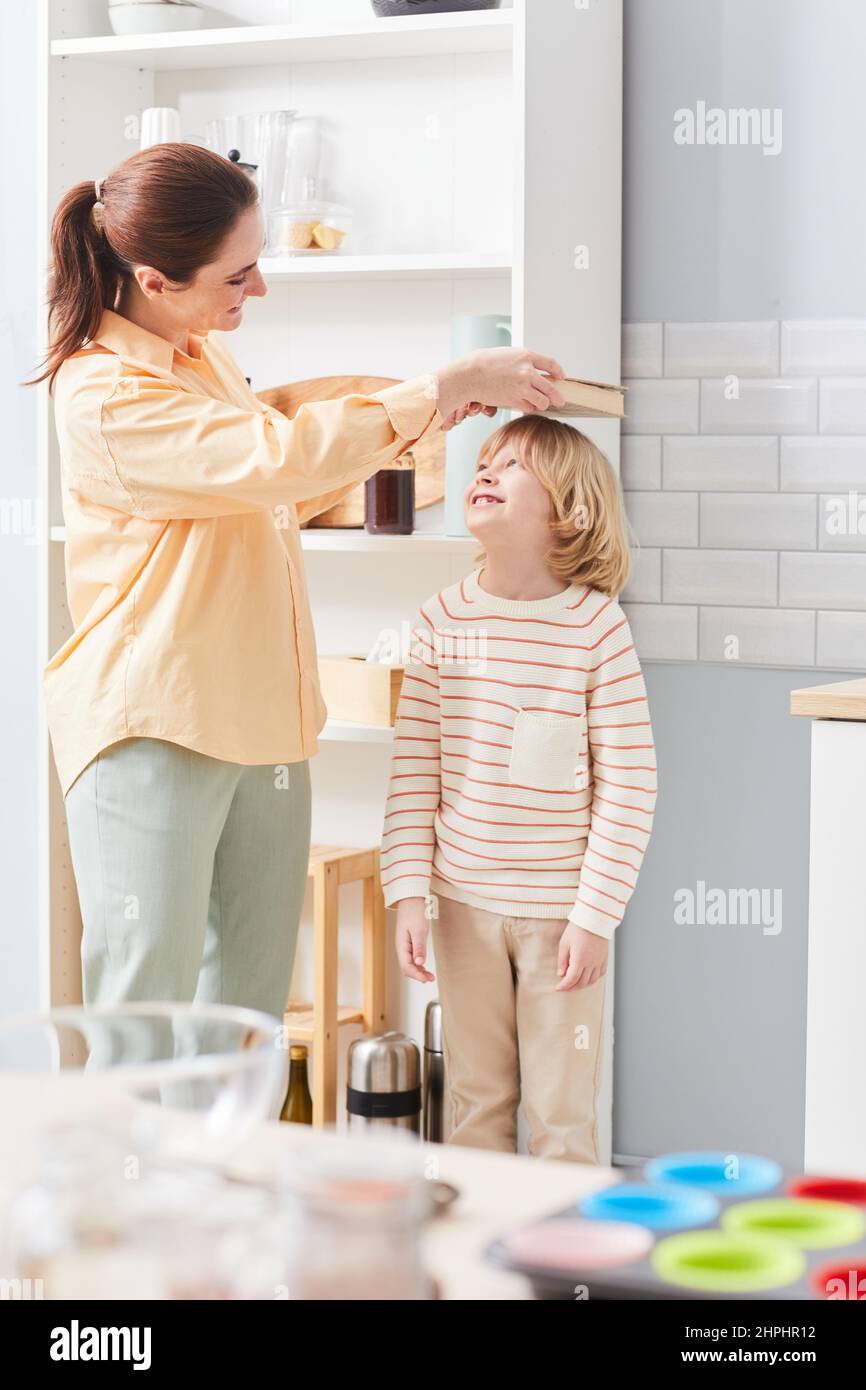 Vertical portrait of happy young mother measuring height of growing boy against kitchen cabinets Stock Photo