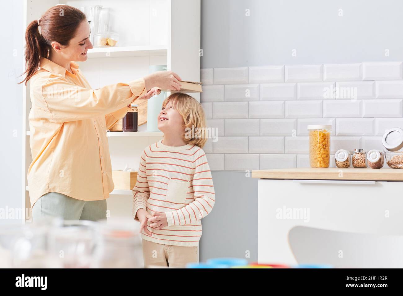 Portrait of happy mother measuring height of growing boy against kitchen cabinets, copy space Stock Photo