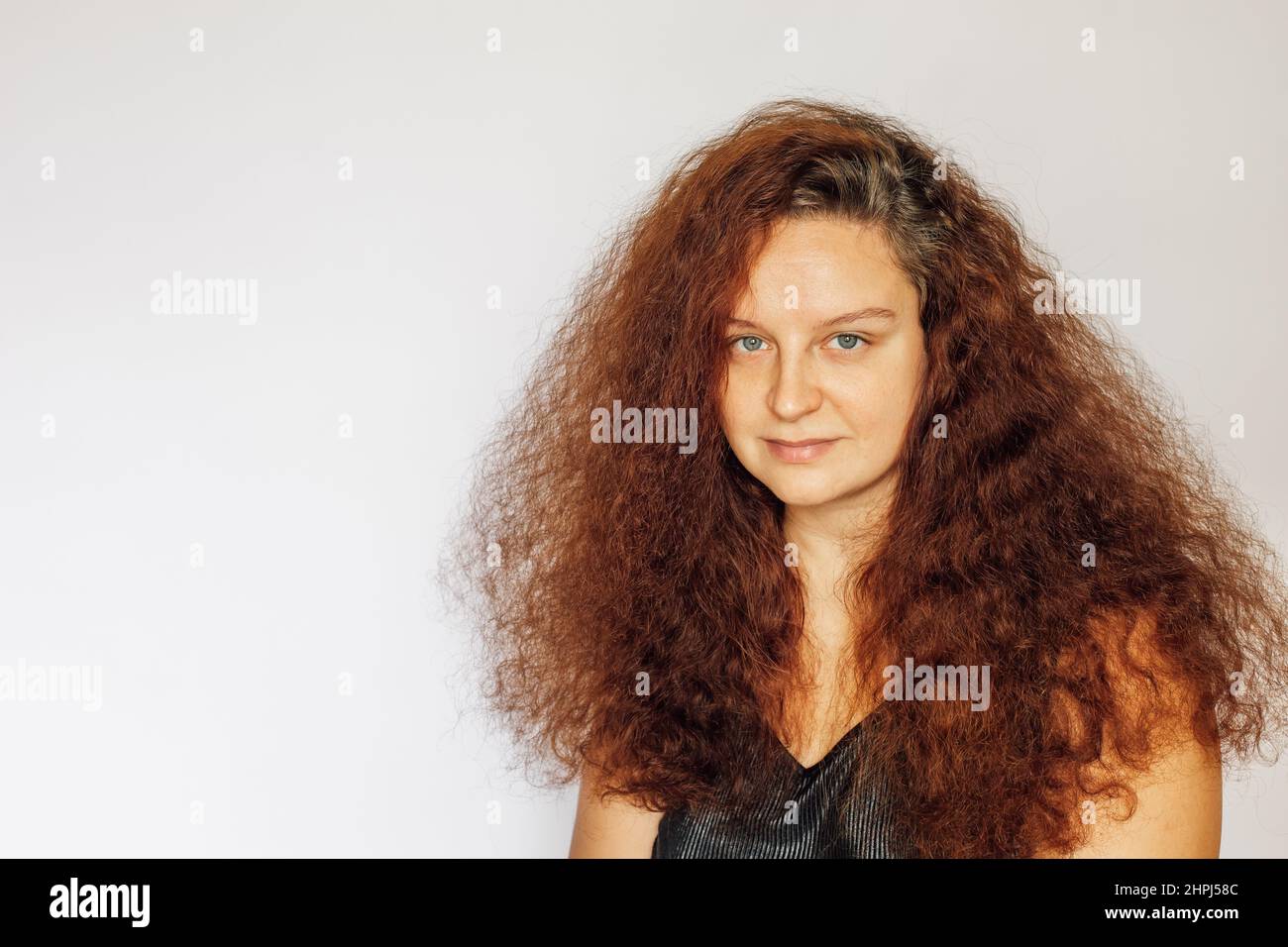 Slightly smiling Caucasian young woman with long curly lush red colored hair, clean face skin and eyes looking at camera on white background. Self Stock Photo