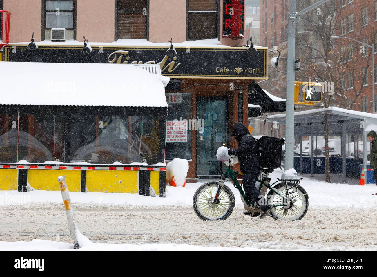 A food delivery bike messenger on an e bike on a snowy, slushy street in New York with an out of business, vacant restaurant for rent in the backgrnd Stock Photo