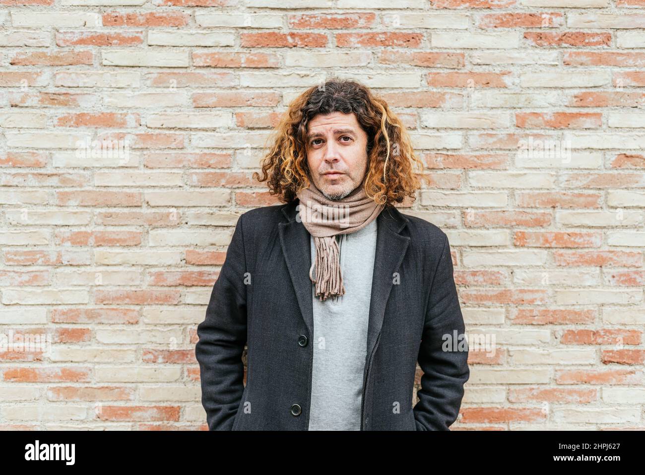 Middle aged male in smart casual coat with scarf and curly hair holding hands in pockets. He is looking at camera while standing against brick wall on city street Stock Photo