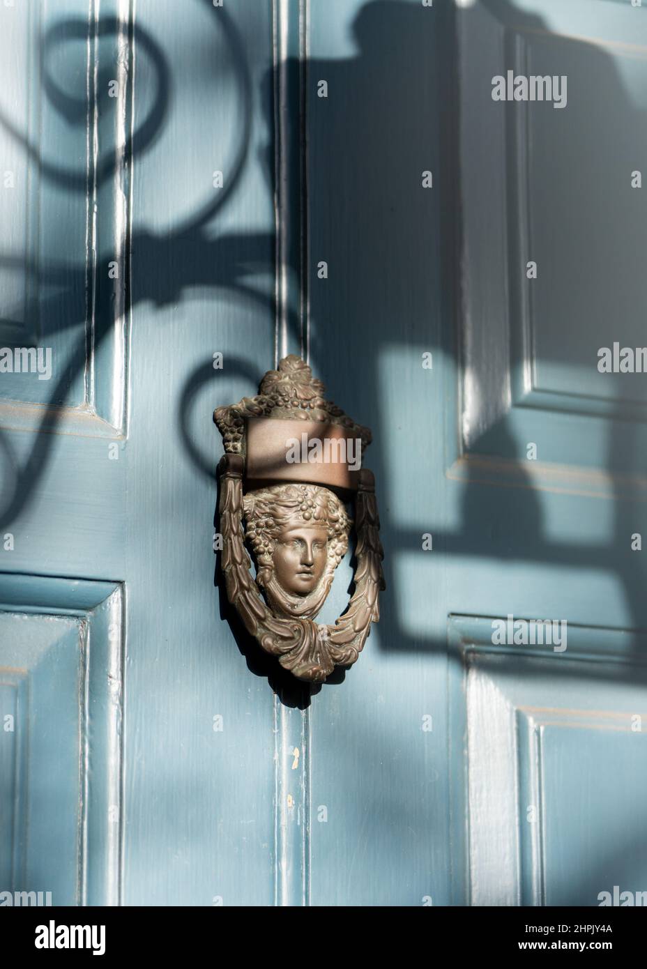Ornate old brass door knocker with face on aqua blue wooden painted door. Shadow cast from street lamp on sunny day. Stock Photo