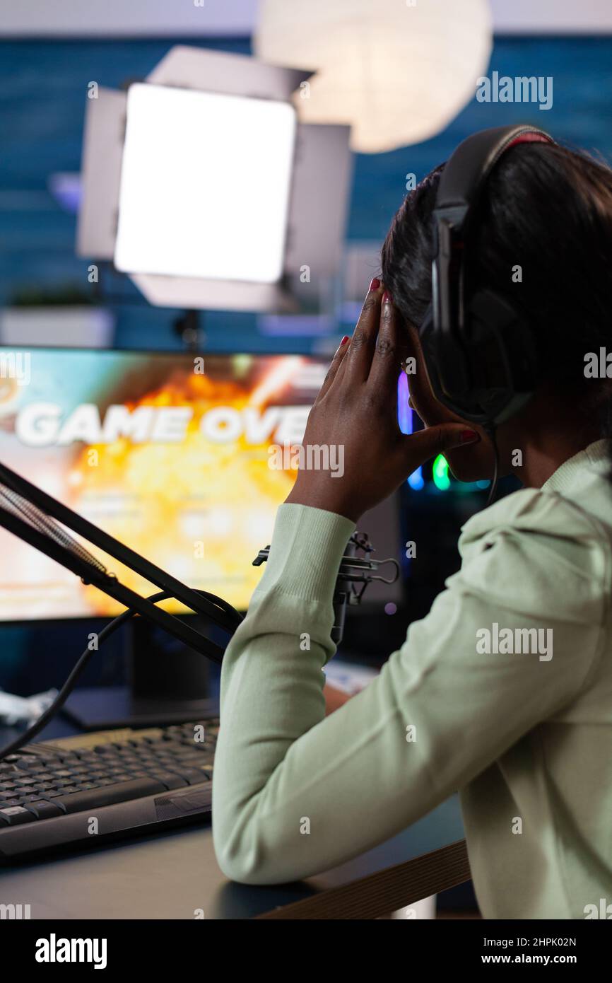 African american woman gamer losing online videogames during space shooter championship. Pro streamer wearing headphones talking with players on live chat playing game using RBG computer. Esport games Stock Photo