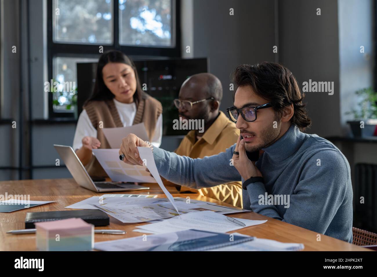 Young busy economist with mobile phone by ear looking through data in financial document against two colleagues discussing papers Stock Photo