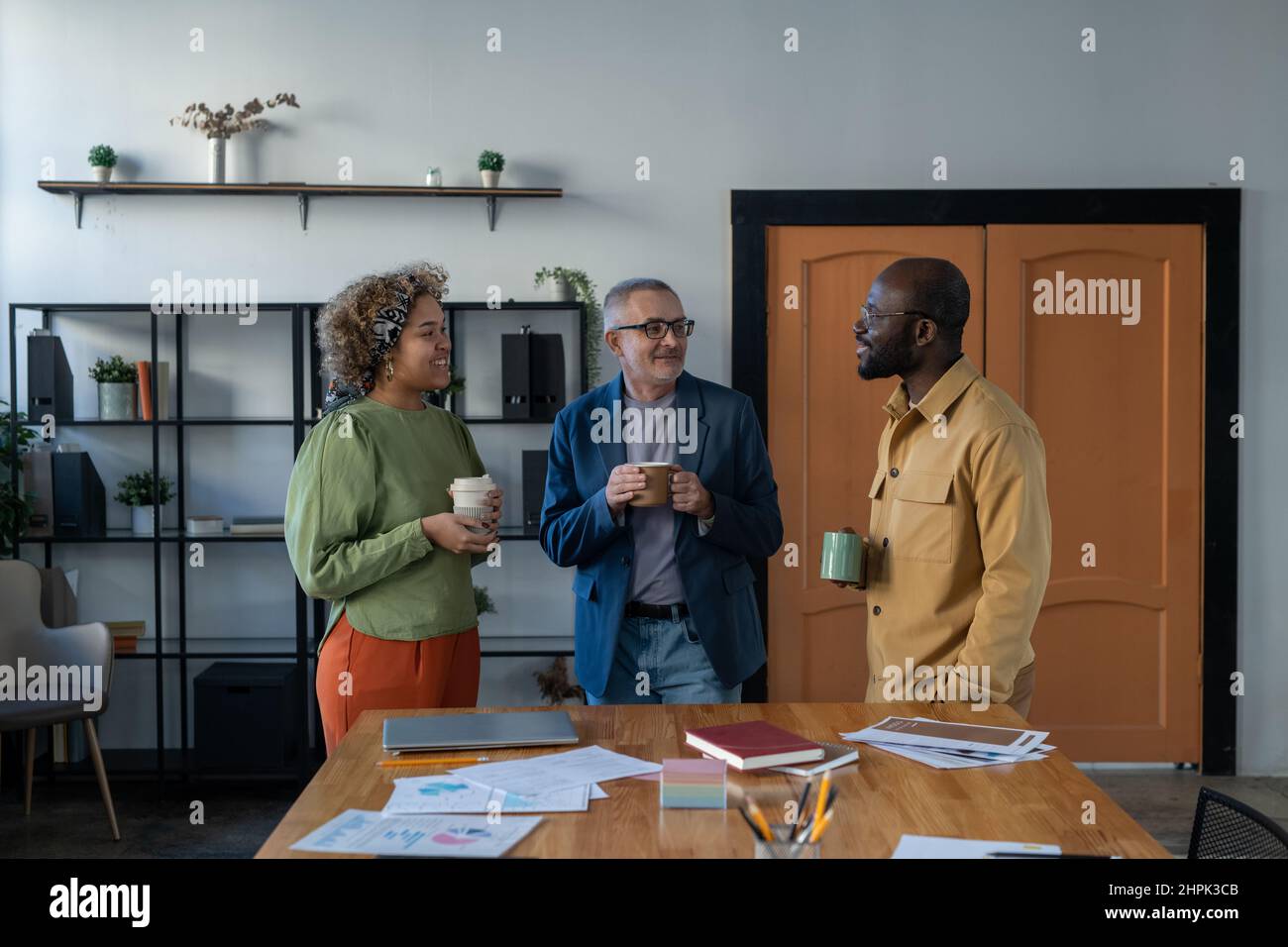 Three confident intercultural colleagues having drinks and talking at break while standing by large table with financial documents in office Stock Photo