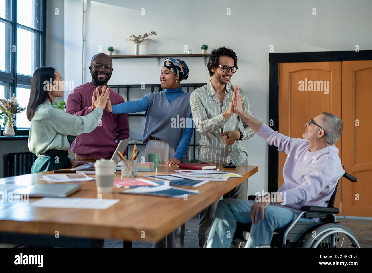Successful intercultural colleagues giving each other high five as symbol of team building while standing by table with financial papers Stock Photo