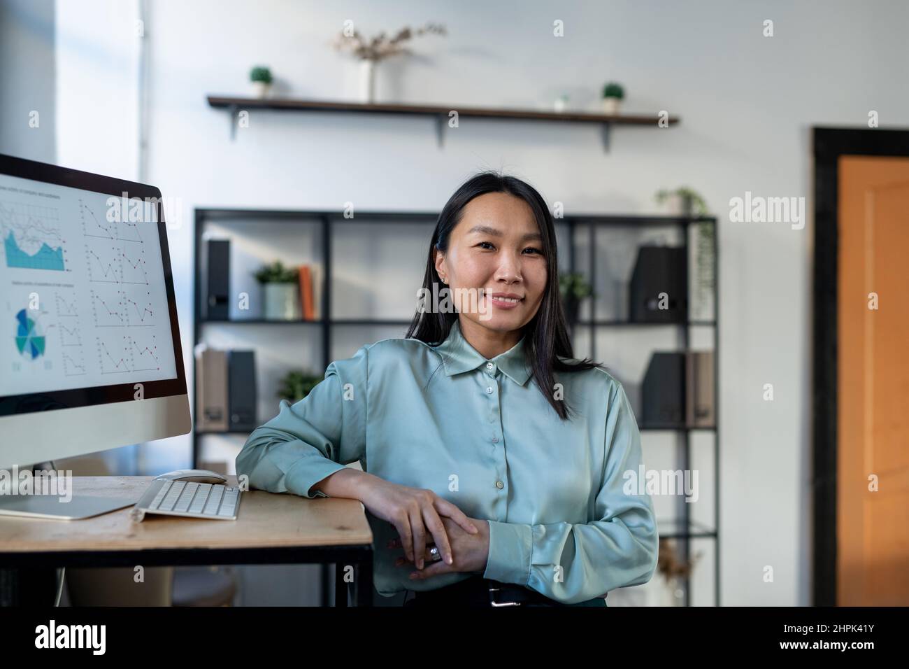 Young Asian female economist in smart casualwear standing by workplace with computer monitor and financial data on screen Stock Photo