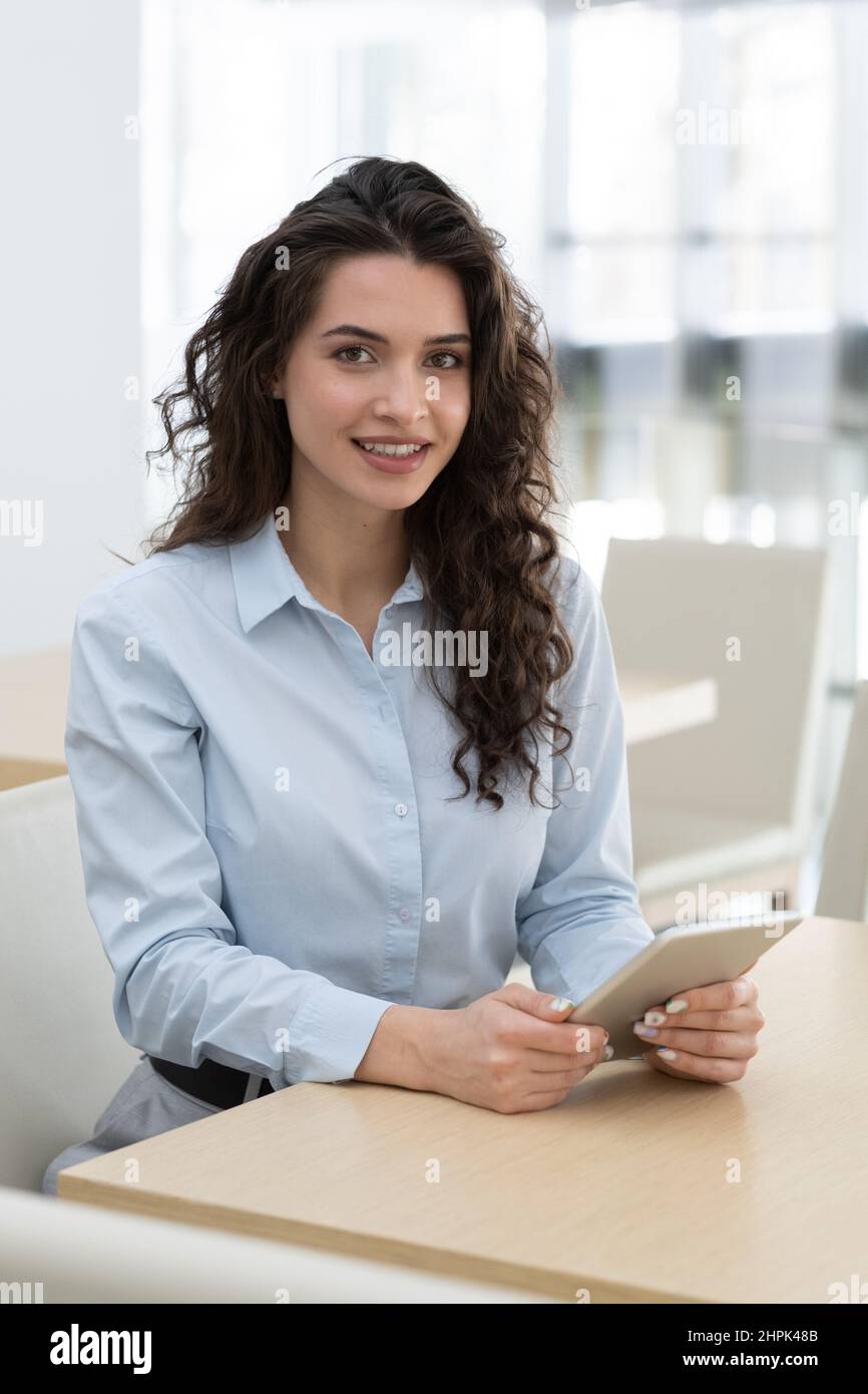 Young pretty owner of high tech business with digital tablet sitting by workplace in modern office and looking at camera Stock Photo