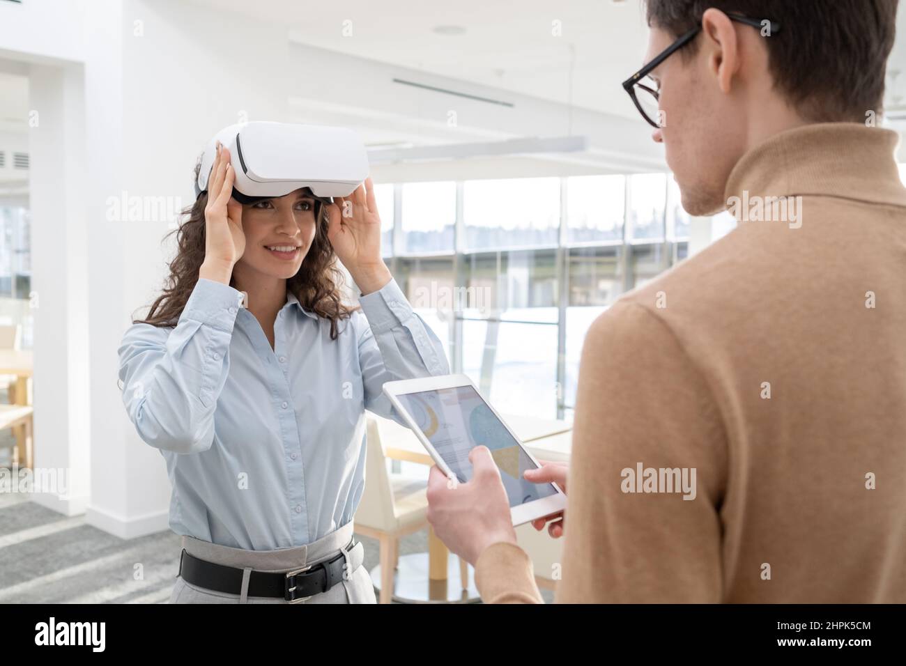 Happy young female employee touching vr headset while communicating with male colleague working with online data in digital tablet Stock Photo