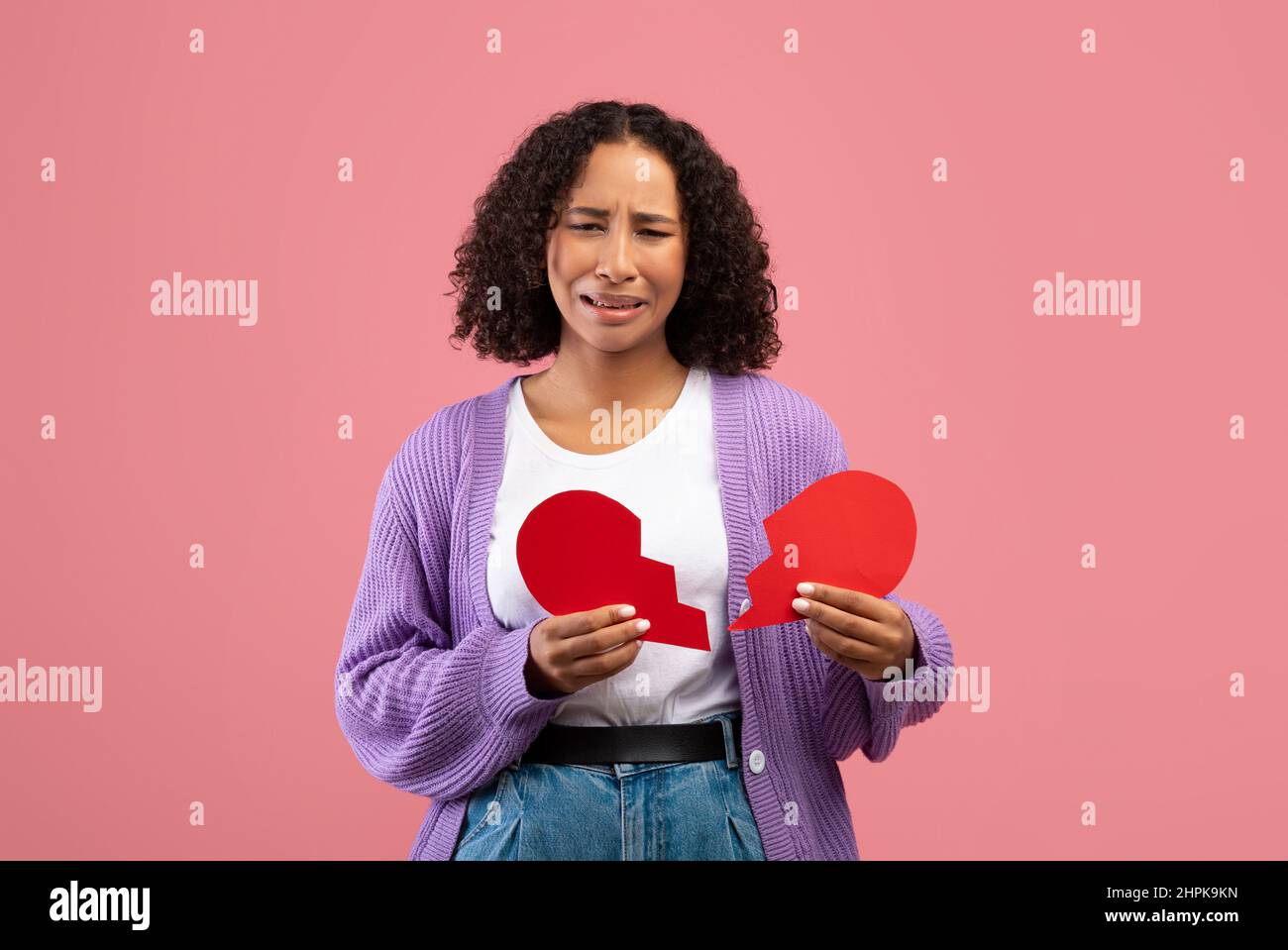 Unhappy young black woman holding two halves of cut paper heart and crying on pink studio background Stock Photo