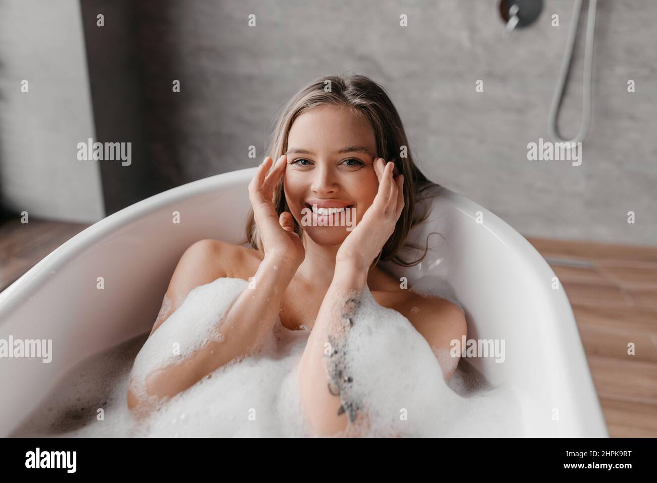 Skin care and spa treatments at home. Happy relaxed lady in bathtub with foam, touching her smooth face skin and smiling Stock Photo
