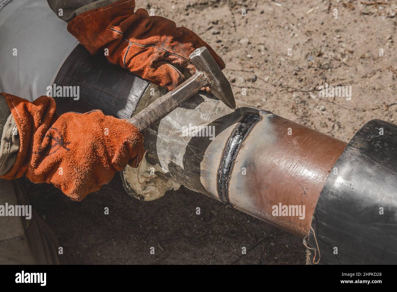 Welder with a hammer in hand and working mittens removes slag after welding industry work the polyethylene water supply main heating pipe on construct Stock Photo