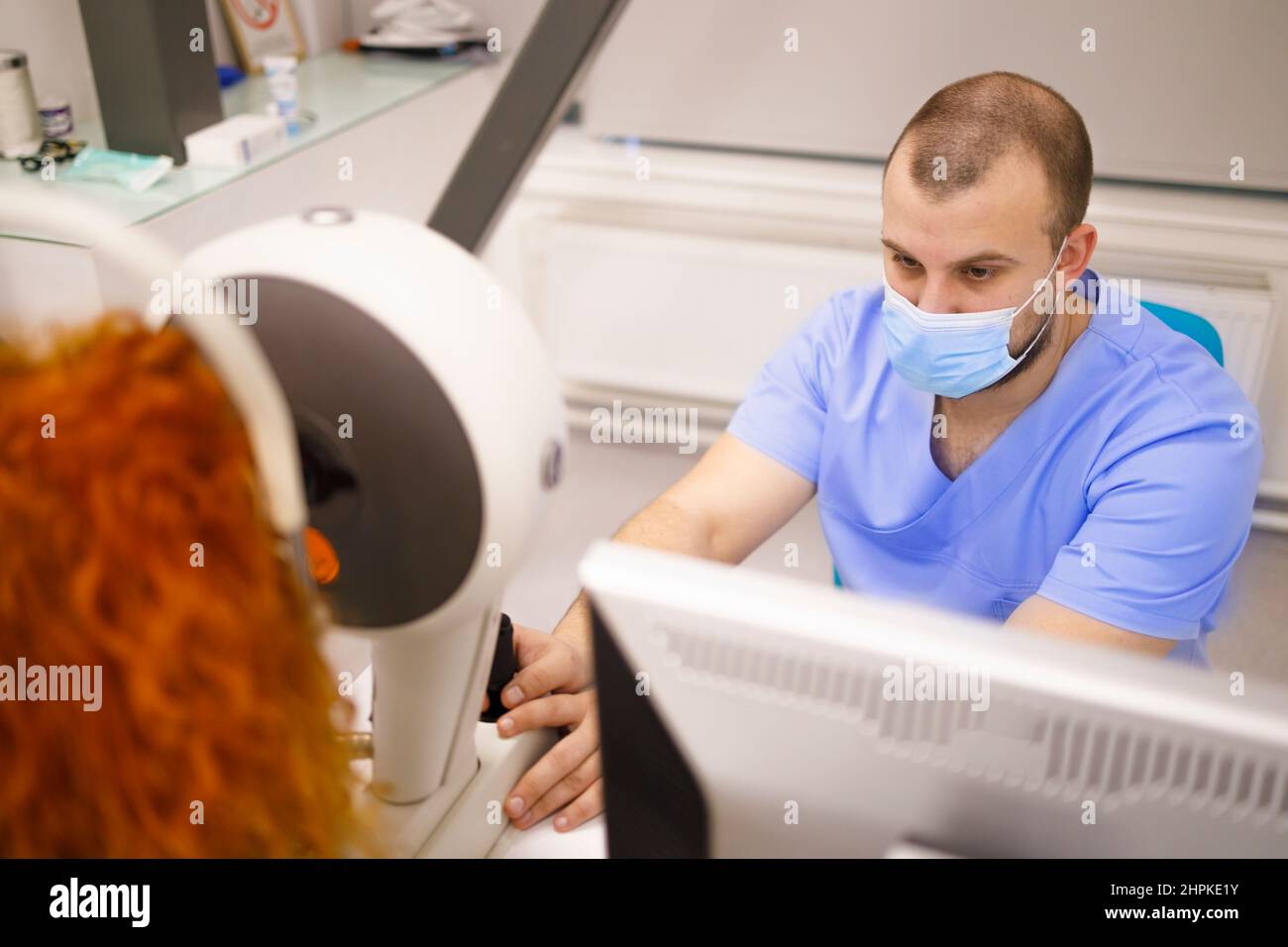 Male technician with a face-mask operating equipment that examines the eyes of a female customer Stock Photo