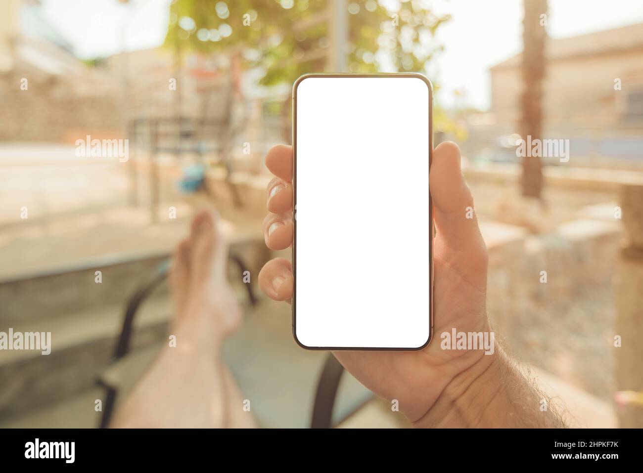 Relaxed man sitting on apartment terrace with feet on the table looking at mobile smart phone mockup screen, selective focus Stock Photo