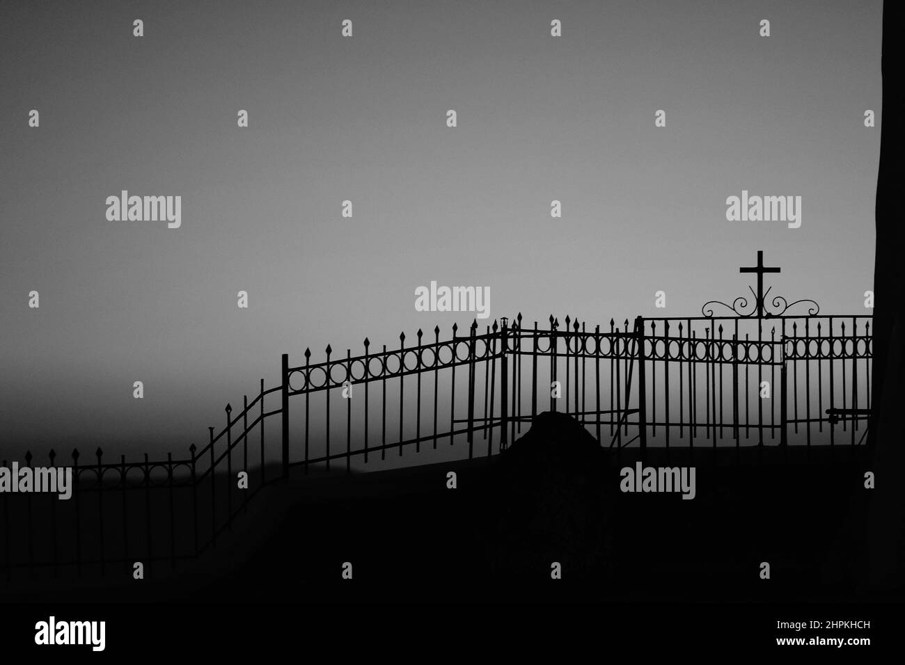 A fence with a religious cross attached to it next to a church and the aegean sea, while the sun is setting in a dramatic way in black and white Stock Photo
