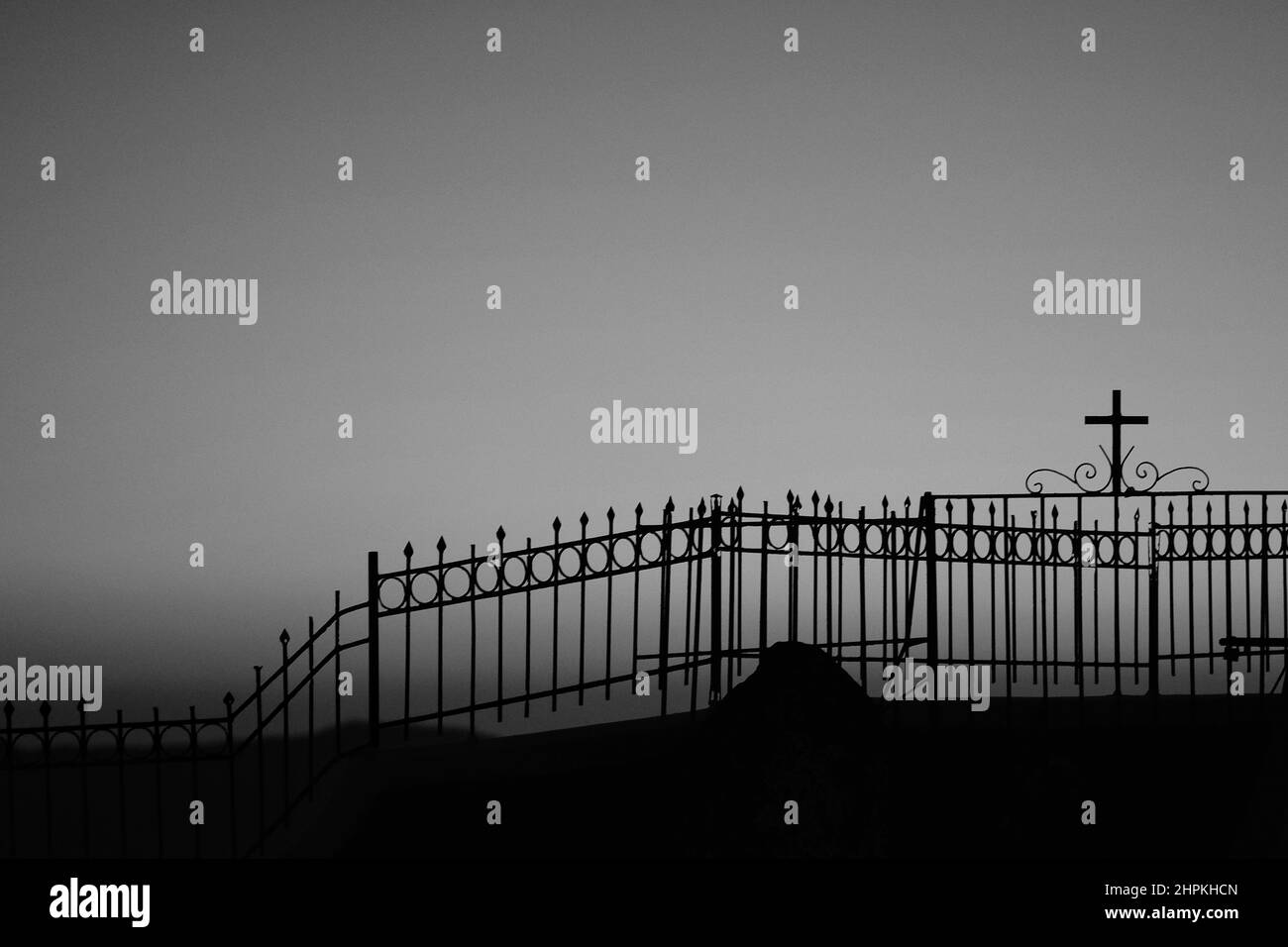 A fence with a religious cross attached to it next to a church and the aegean sea, while the sun is setting in a dramatic way in black and white Stock Photo