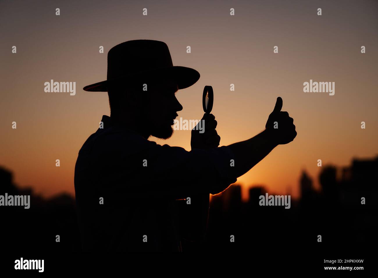 Male silhouette in hat looking with magnifying glass showing thumbs up gesture. Man searching sales or discounts at sunset using loupe with sun on background and urban view. Investigation concept Stock Photo