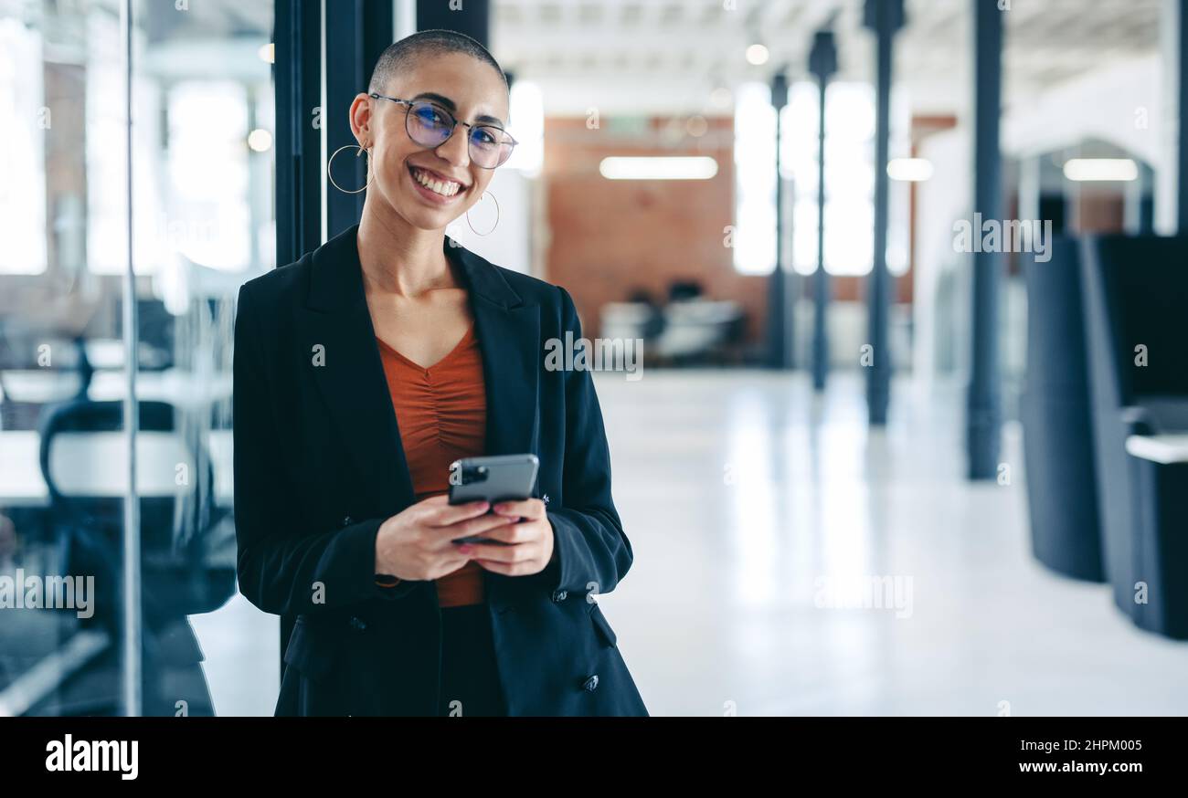 Confident businesswoman smiling at the camera while holding a smartphone. Happy young businesswoman standing alone in a modern workplace. Young busine Stock Photo