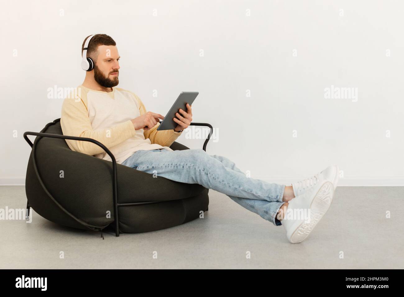 Serious Guy Using Tablet Wearing Earphones Sitting Indoor, Side View Stock Photo