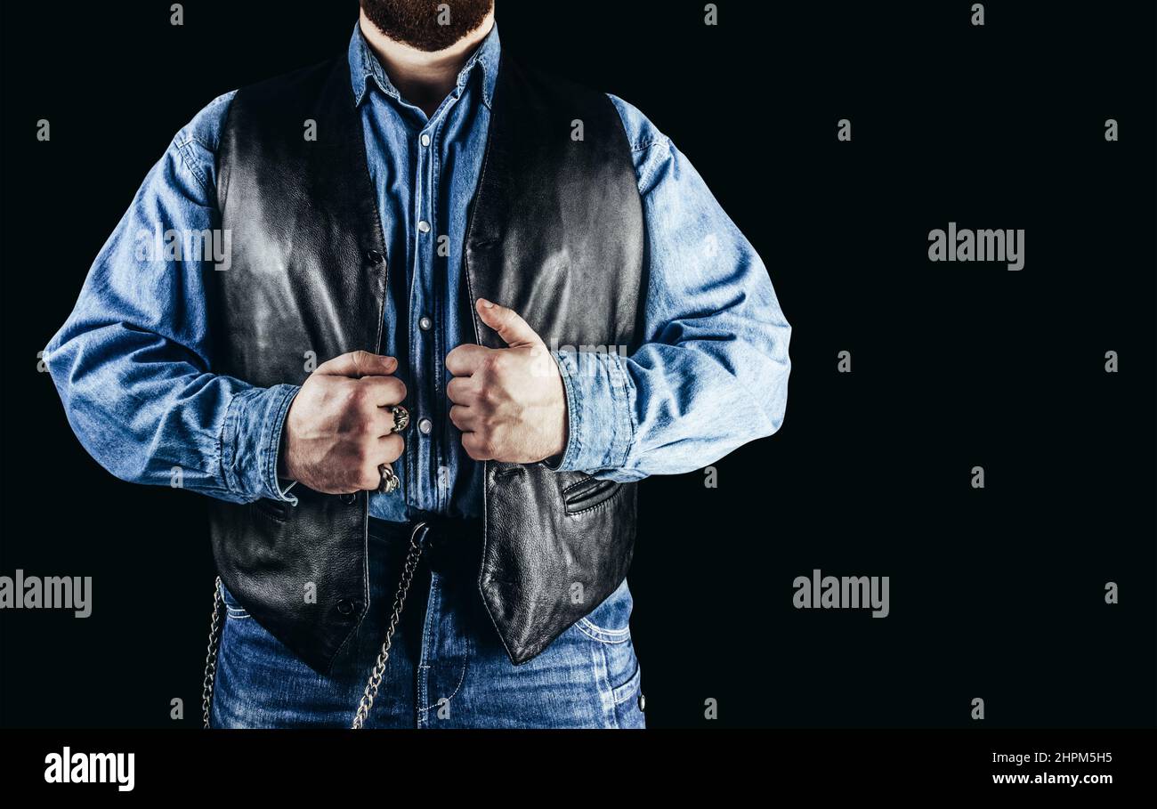 Front view photo of man in jeans shirt, denim pants and leather biker vest standing on black background. Stock Photo