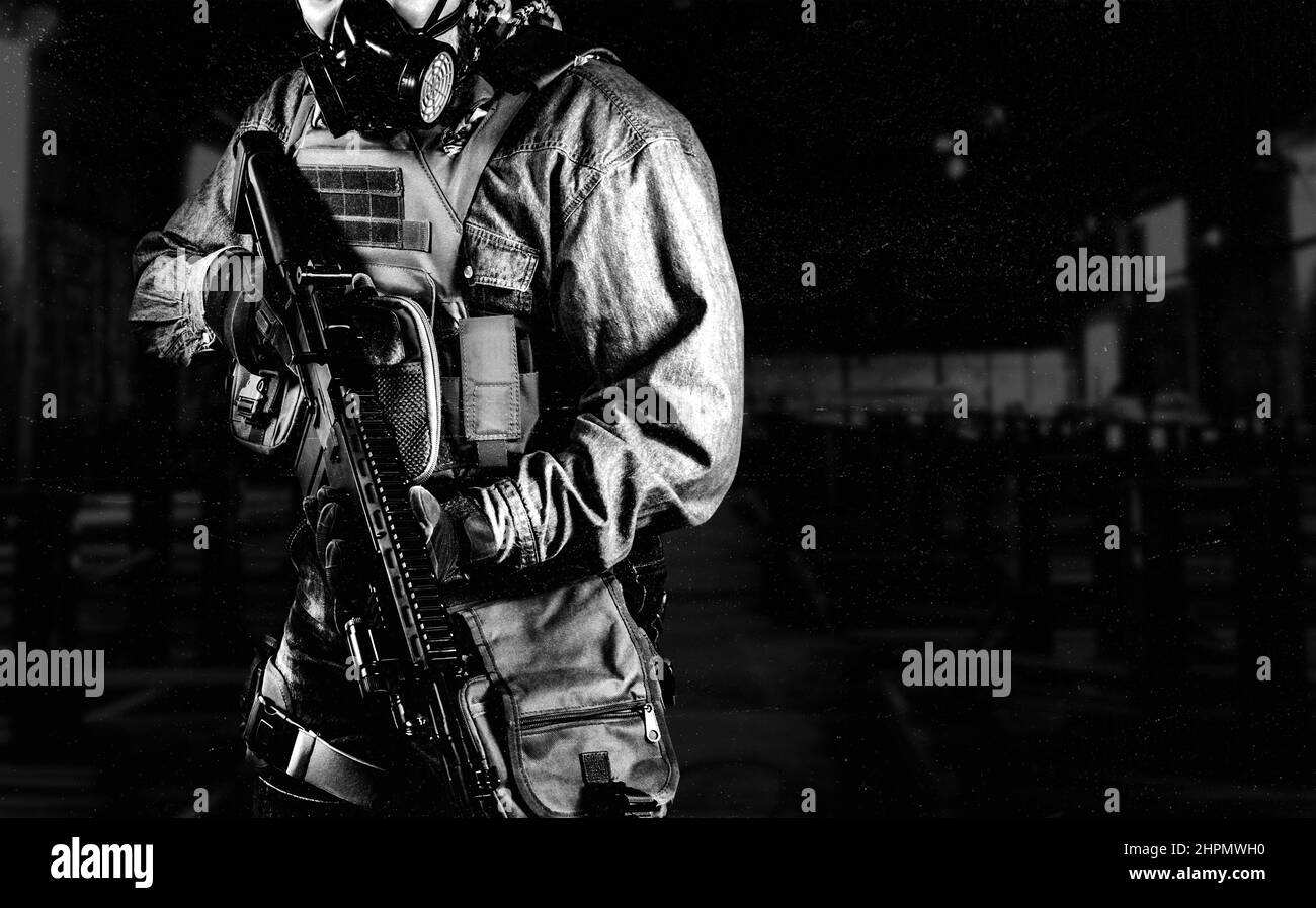 Black and white photo of urban soldier in tactical military outfit and gas mask standing with rifle and gas mask on dark factory background. Stock Photo