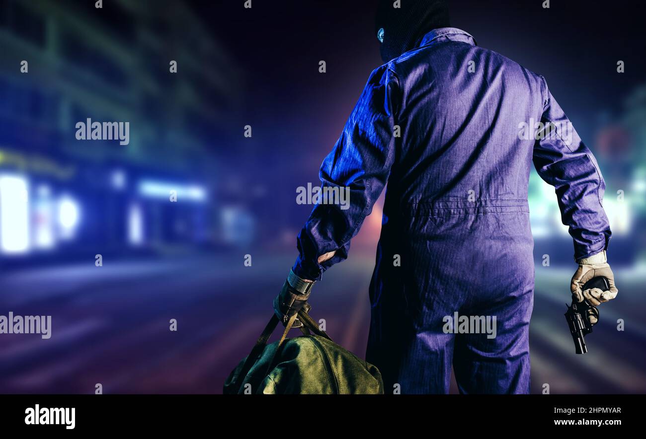 Photo of robber in mask, overalls, gloves, bag and gun standing back view on night street background. Stock Photo