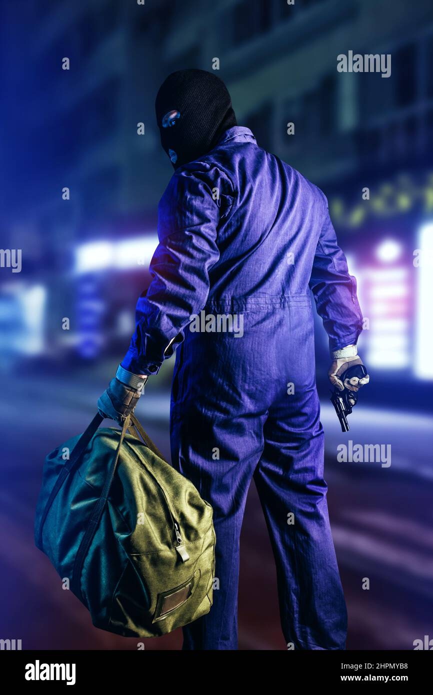 Photo of robber in mask, overalls, gloves, bag and gun standing back view on night street background, vertical oriented. Stock Photo