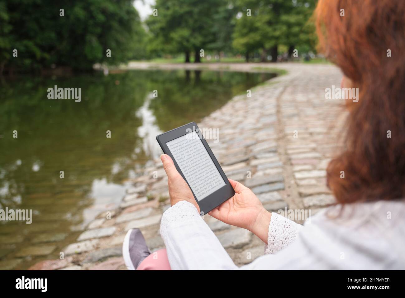 Unrecognizable mature woman reading an ebook on an electronic reader sitting in a park. Concepts: technology and reading. Image with copy space Stock Photo