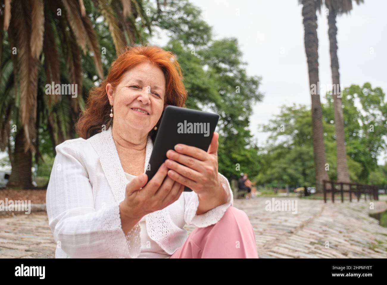 Mature hispanic woman reading an ebook on an electronic reader sitting in a park. Concepts: technology and reading. Image with copy space Stock Photo