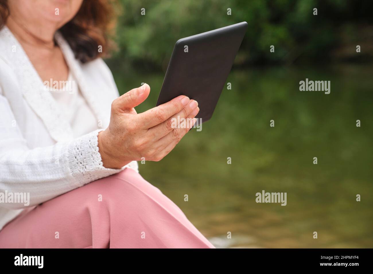 Unrecognizable mature woman reading an ebook on an electronic reader sitting by a lake in a park. Concepts: technology and reading. Image with copy sp Stock Photo
