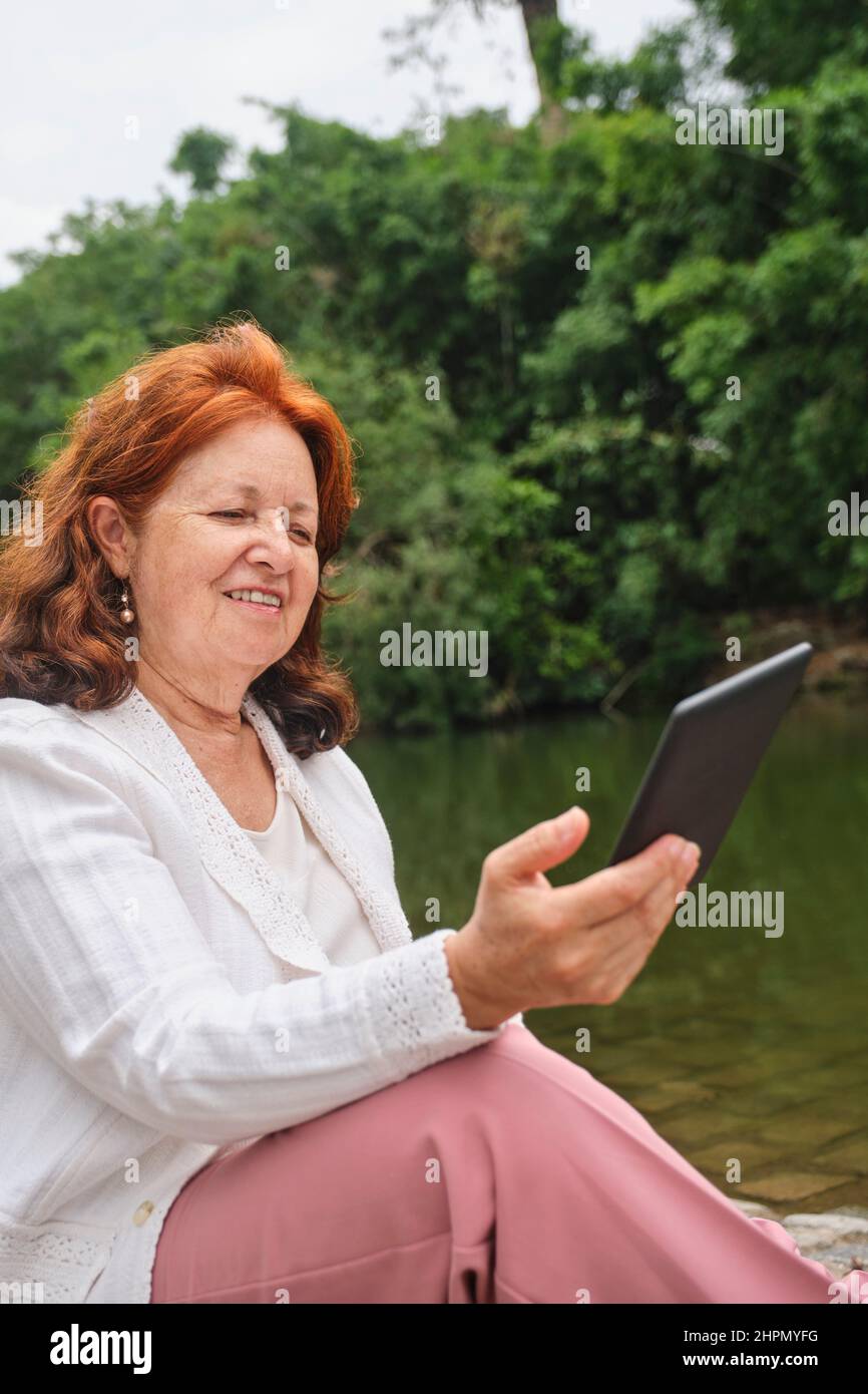 Mature hispanic woman reading an ebook on an electronic reader sitting by a lake in a park. Concepts: technology, reading and relaxing outdoors. Verti Stock Photo