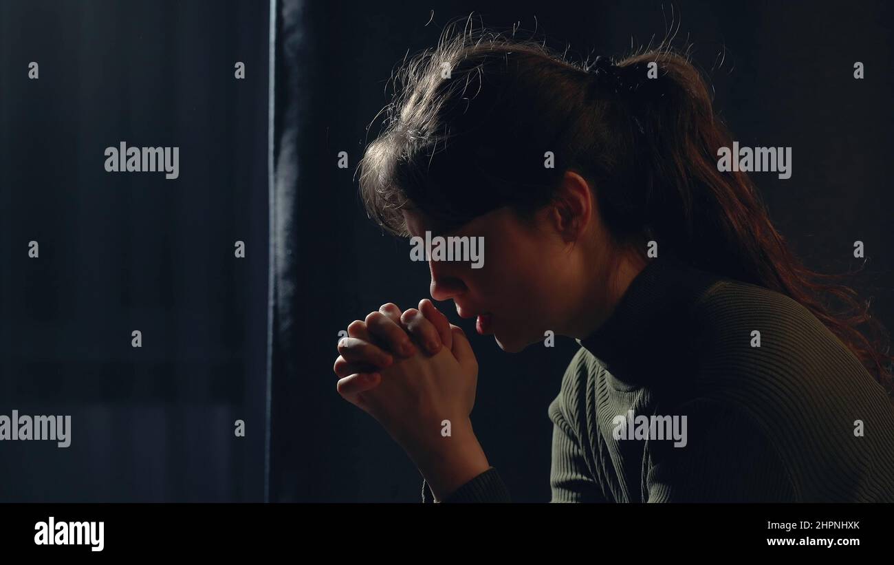 Close up portrait of young woman praying at home in dark room. Girl crossed her fingers, bowed head and prays to god. Faith and hope concept. Stock Photo
