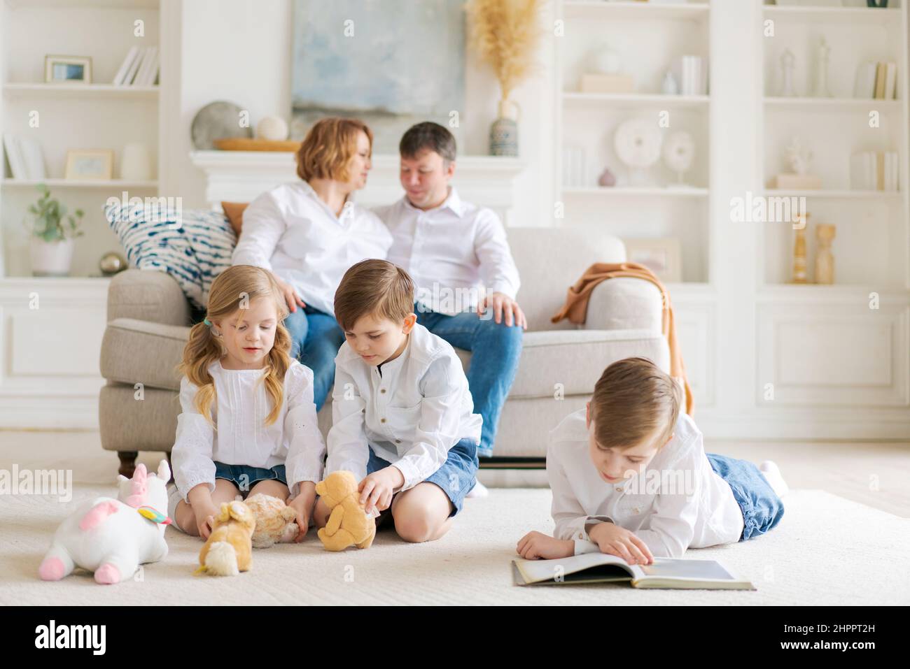 Happy caucasian parents are relaxing on cozy sofa in living room at home. Small children playing on a warm floor and reading a book. An overjoyed family rests on the weekend in their own home. Stock Photo
