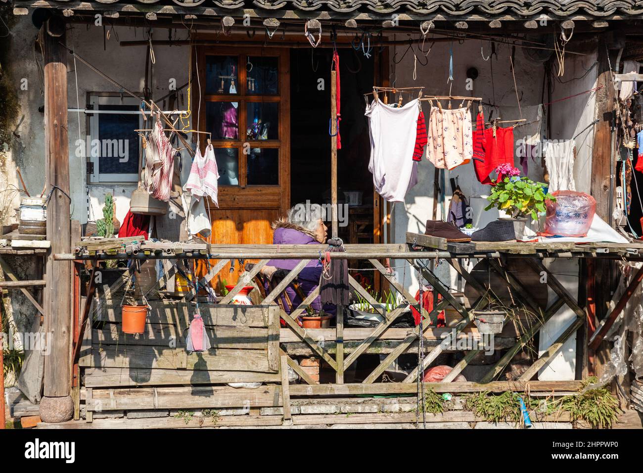 Woman sitting on a balcony cluttered with various objects, trying to warm himself in the winter sun in the water village of Wenzhou, China Stock Photo