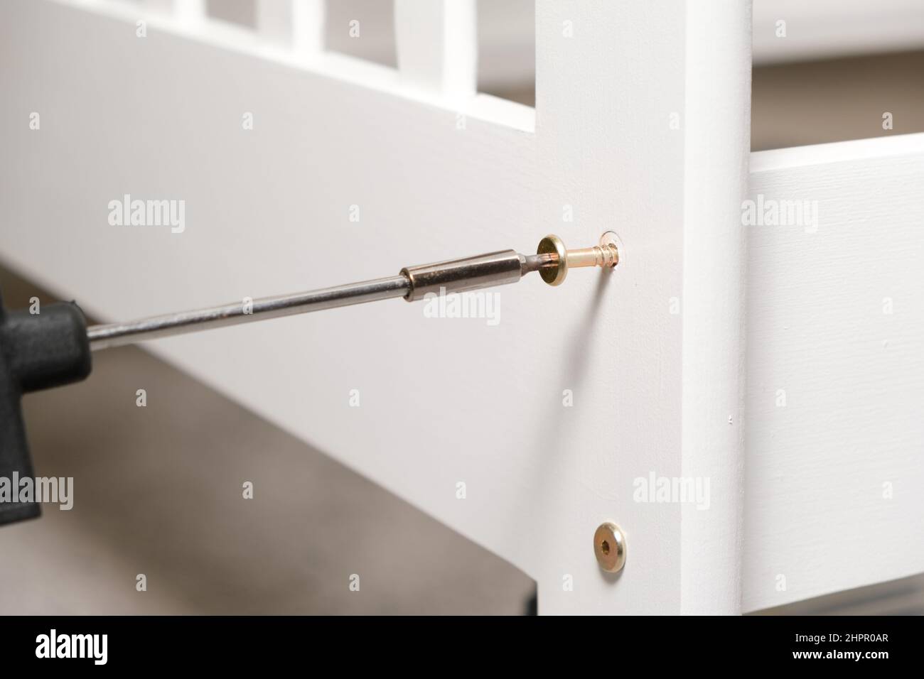 Close-up process of screwing a self-tapping screw into a white wooden bed with a screwdriver. Stock Photo