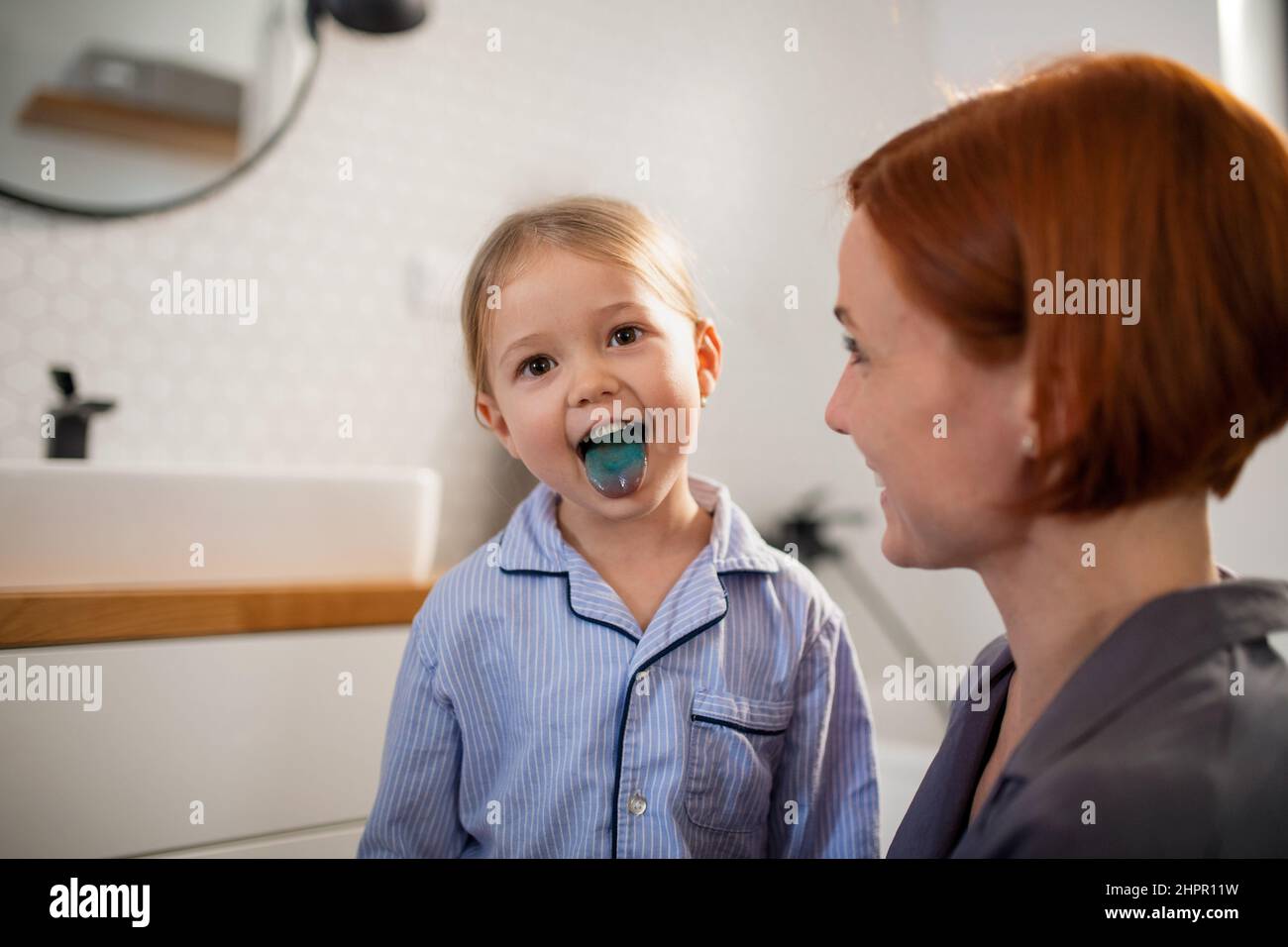 Little girl sticking showing her blue tongue to mother in bathroom at home. Stock Photo