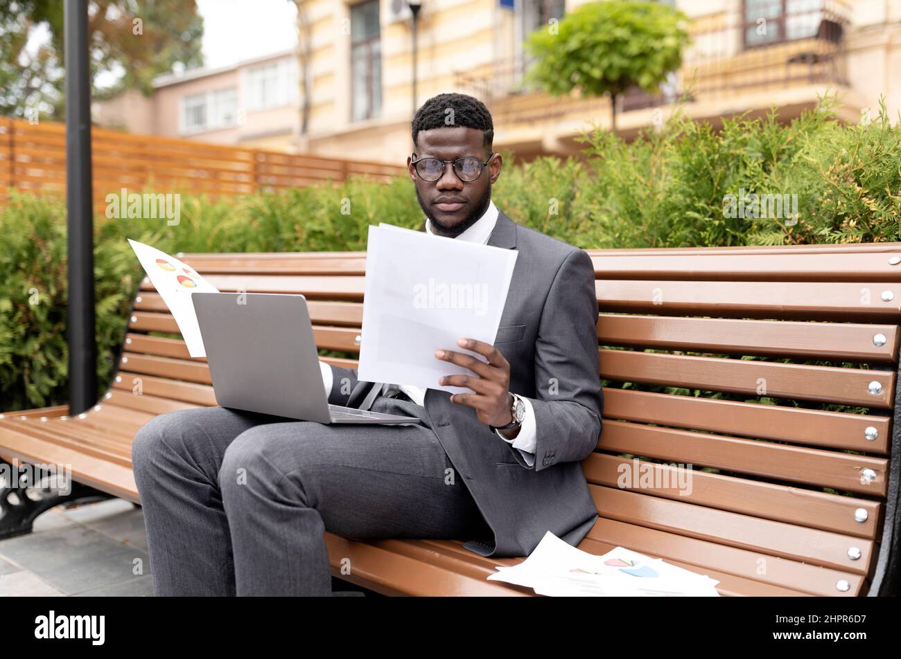 Business career concept. Serious african american entrepreneur working on laptop and reading documents, sitting on bench Stock Photo