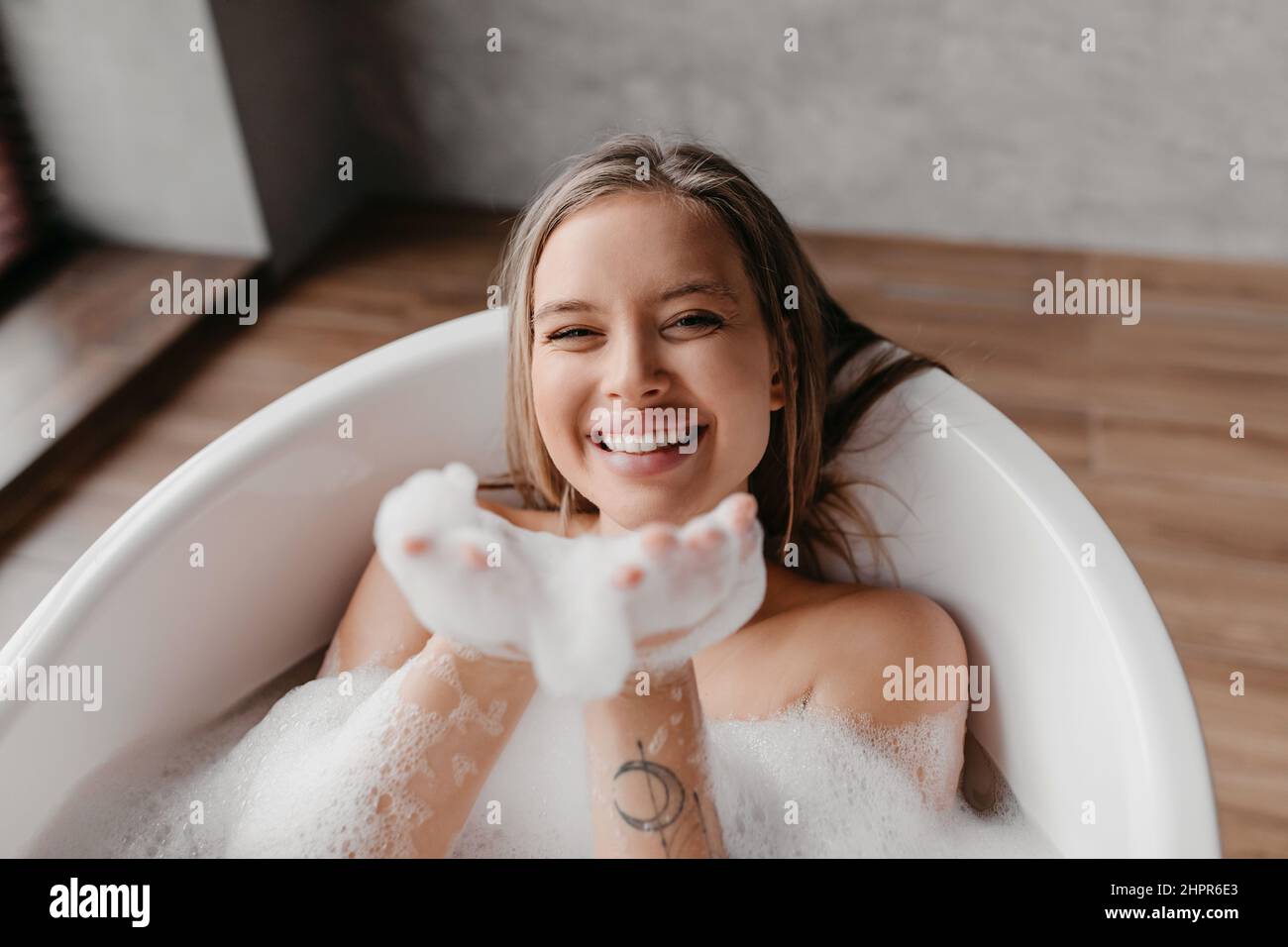 Playful lady having fun while lying in bathtub full of foam, blowing