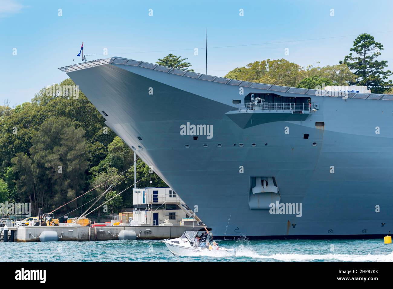 A small leisure boat passes the bow of the Canberra class, Royal Australian Navy landing helicopter dock ship HMAS Adelaide (L01) at Garden Island Stock Photo