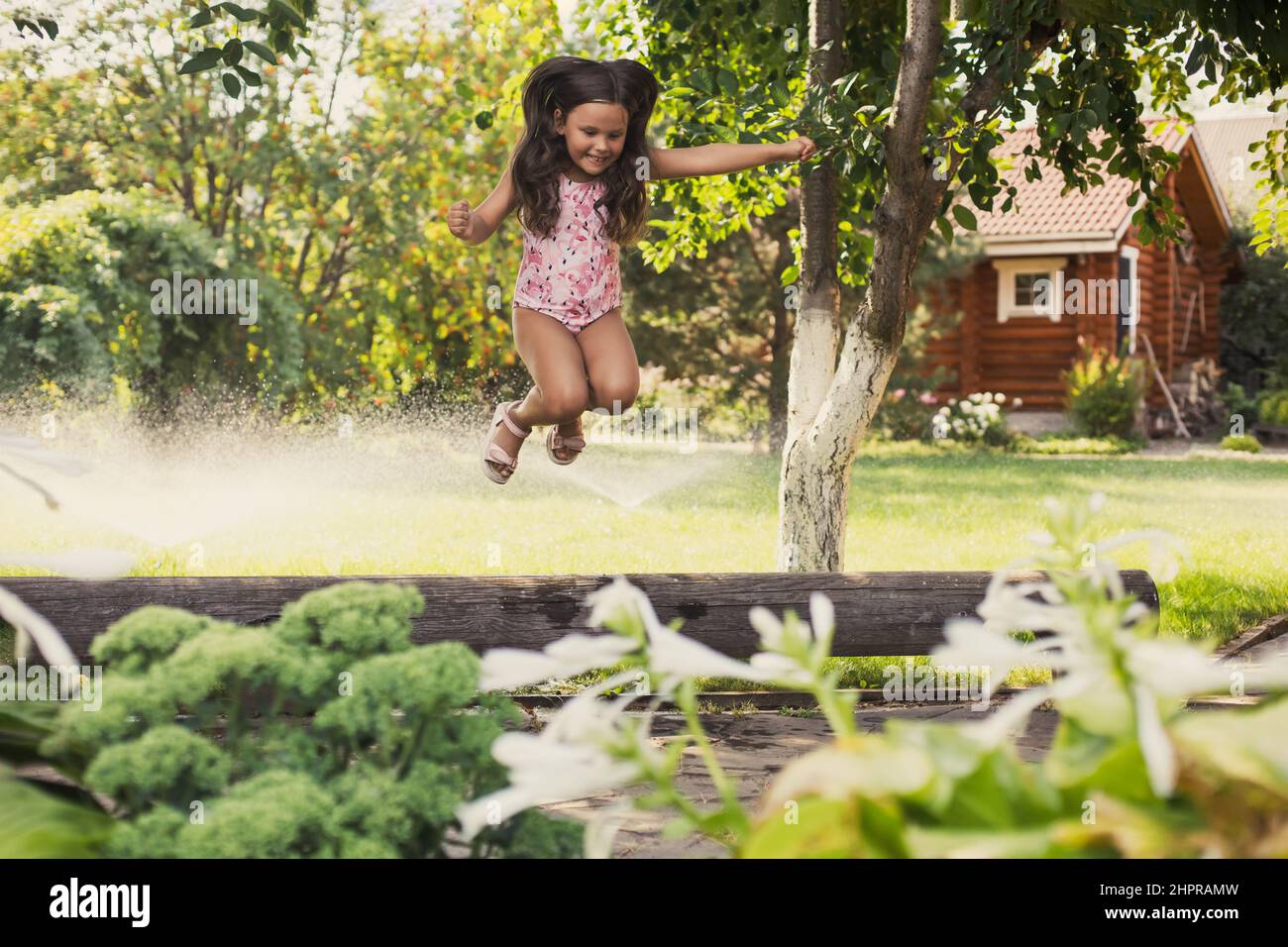 Excited female child playing outside jumping off log looking down in backyard indulging with trees and water sprinklers in background in daytime Stock Photo
