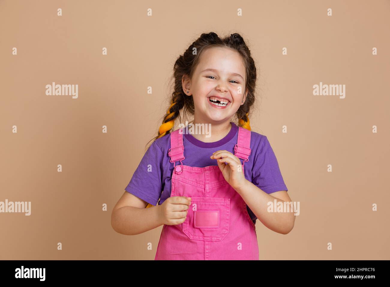 Portrait of laughing small female with kanekalon braids with missing tooth playing, looking at camera wearing pink jumpsuit and purple t-shirt on Stock Photo