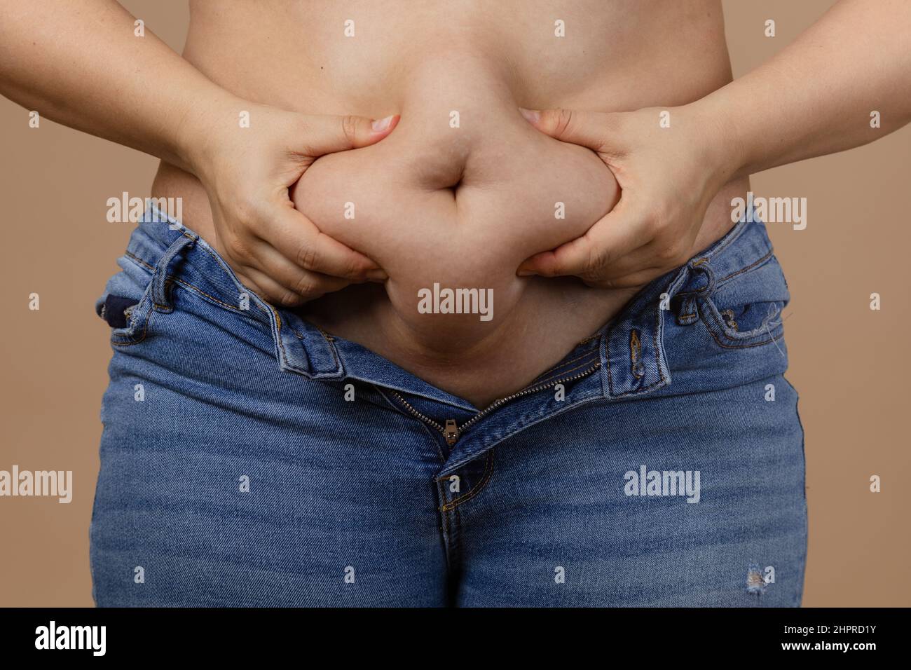 Woman squeezing showing fat sagging tummy in blue unzipped jeans on beige background. Sudden weight gain. Visceral fat. Body positive. Tight little Stock Photo