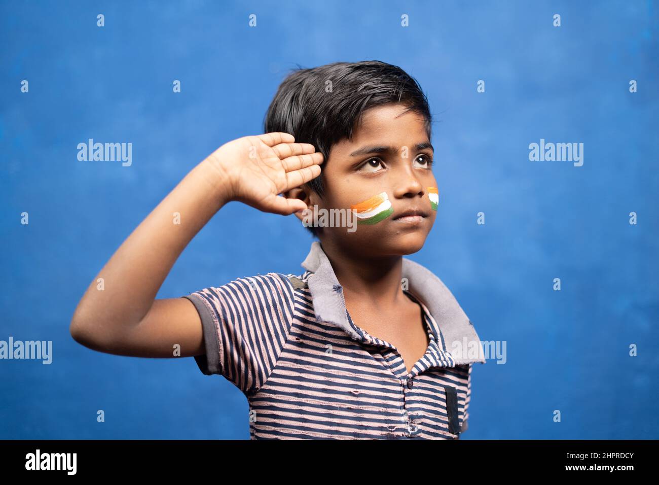 serious kid with torn t-shirt and Indian flag on face saluting by looking above - concept of poverty, patriotism, innocence and independence day. Stock Photo