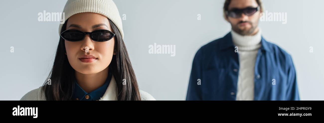 asian woman in white beanie and dark sunglasses looking at camera isolated on grey, banner Stock Photo