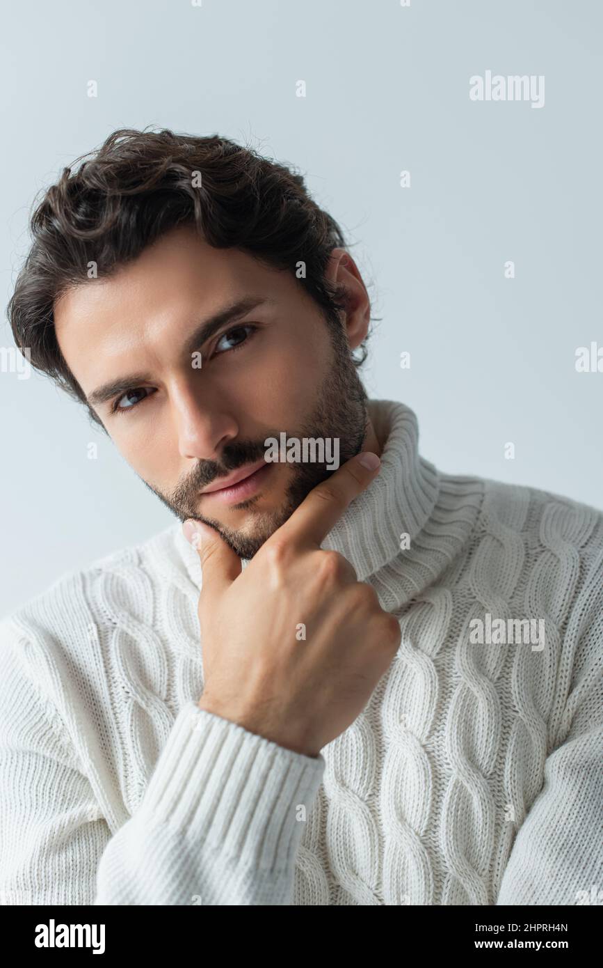 brunette man in white knitted sweater touching chin and looking at camera isolated on grey Stock Photo