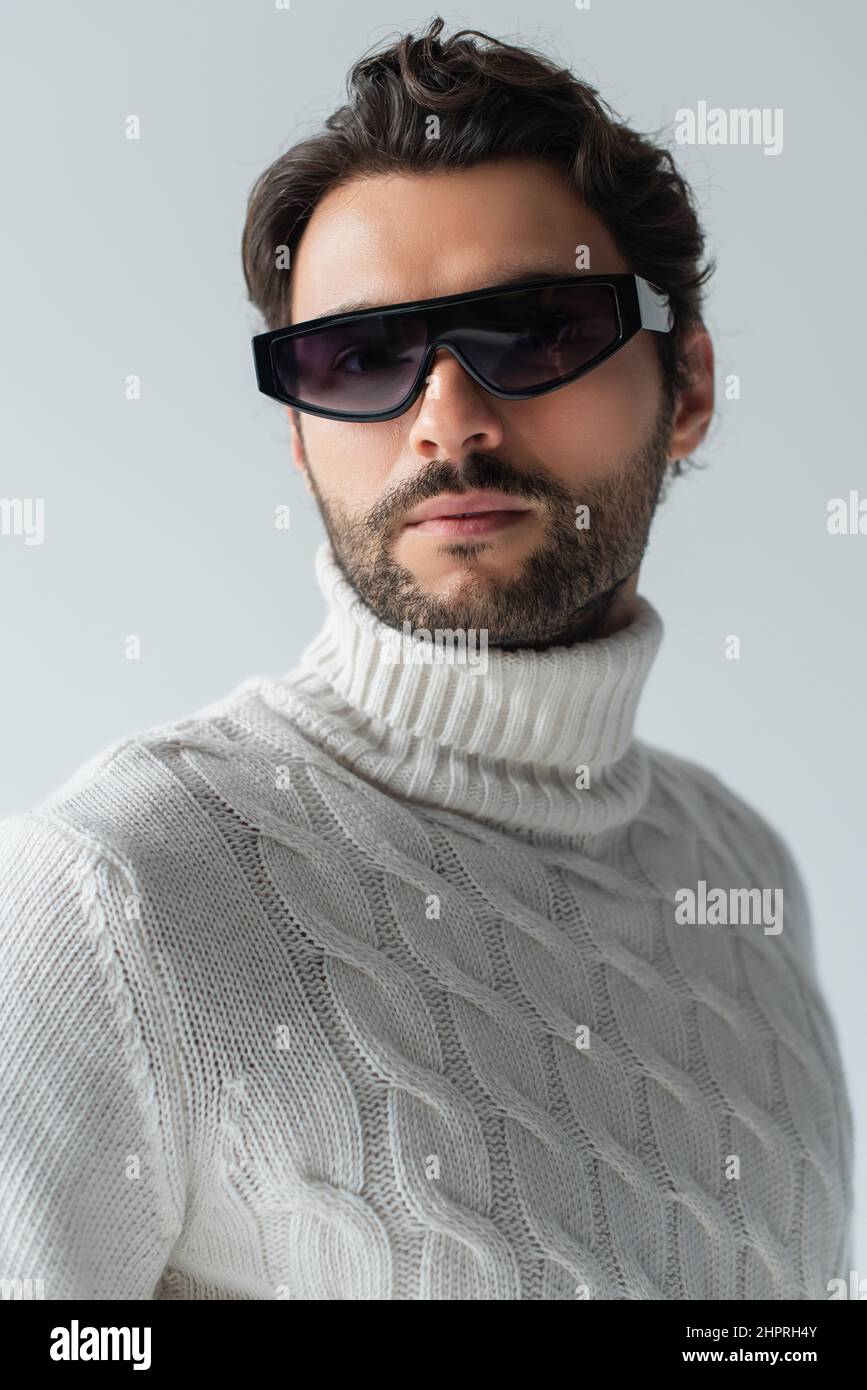 trendy man in white sweater and dark sunglasses looking at camera isolated on grey Stock Photo
