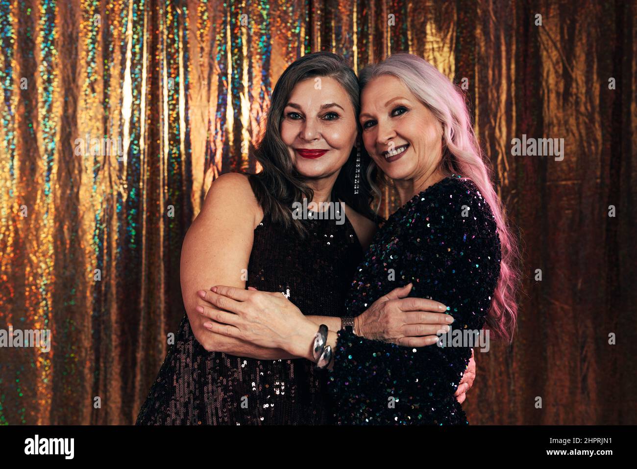 Two smiling senior girlfriends wearing evening dresses hugging each other while standing at glitter background Stock Photo