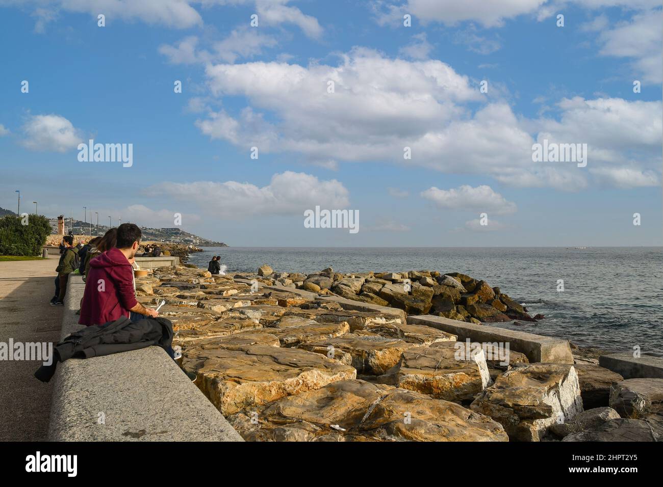 People sitting on the rocky sea shore of Sanremo in the Italian Riviera in a sunny winter day, Imperia, Liguria, Italy Stock Photo