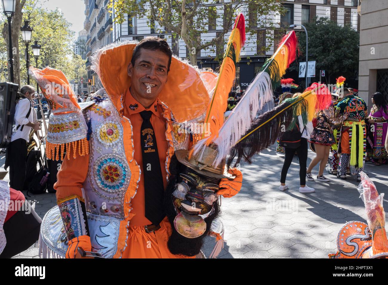 Man wearing a sumptuous white and orange Hispanic-style costume poses proudly holding a traditional Bolivian mask. Hispanic Day in Barcelona, Spain. Stock Photo